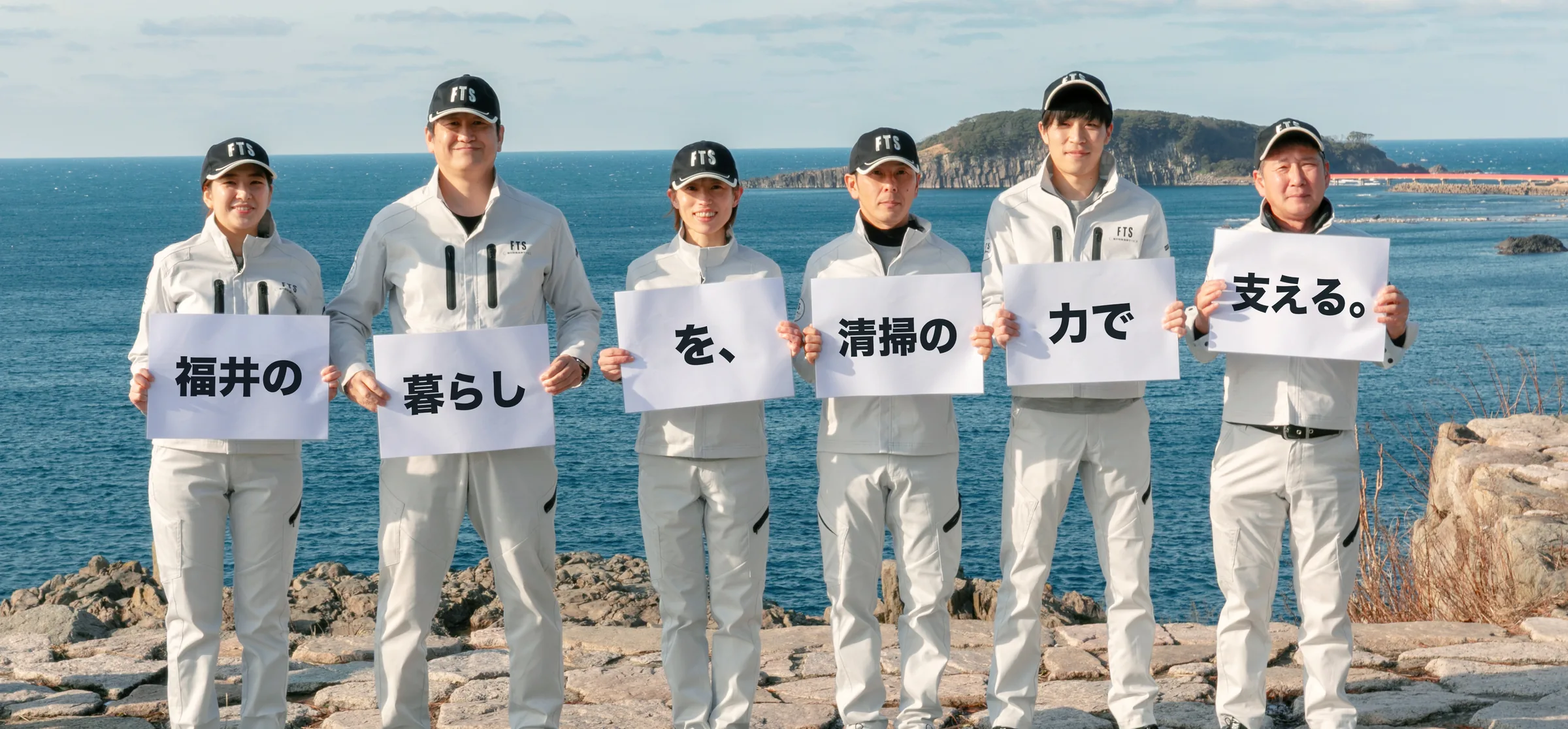 海辺で「福井の暮らしを、清掃の力で支える。」の紙を持つスタッフ